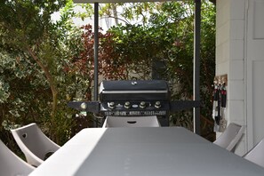 Una parrilla con panel de control, rodeada de sillas blancas, con árboles y una pared blanca de fondo.