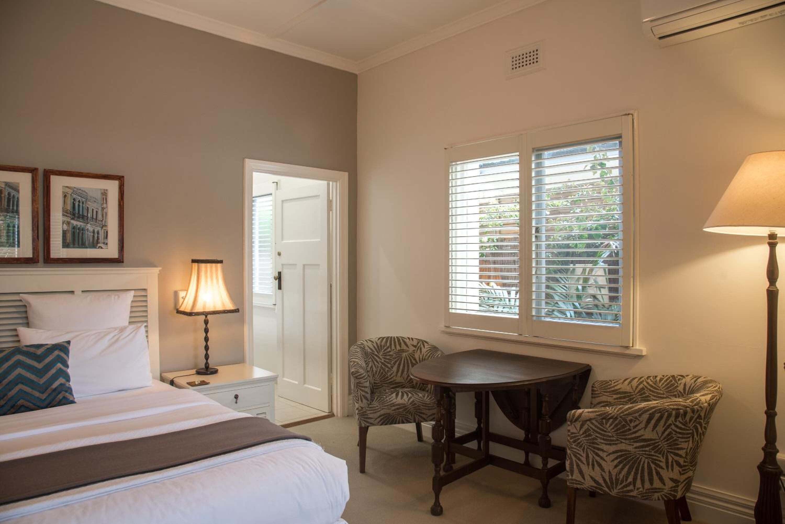 A hotel room with a bed, a bedside table with a lamp, a chair, a table, and a window with blinds.