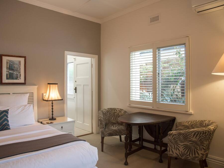 Habitación de hotel con cama, mesita de noche con lámpara, una silla, una mesa y una ventana con persianas.