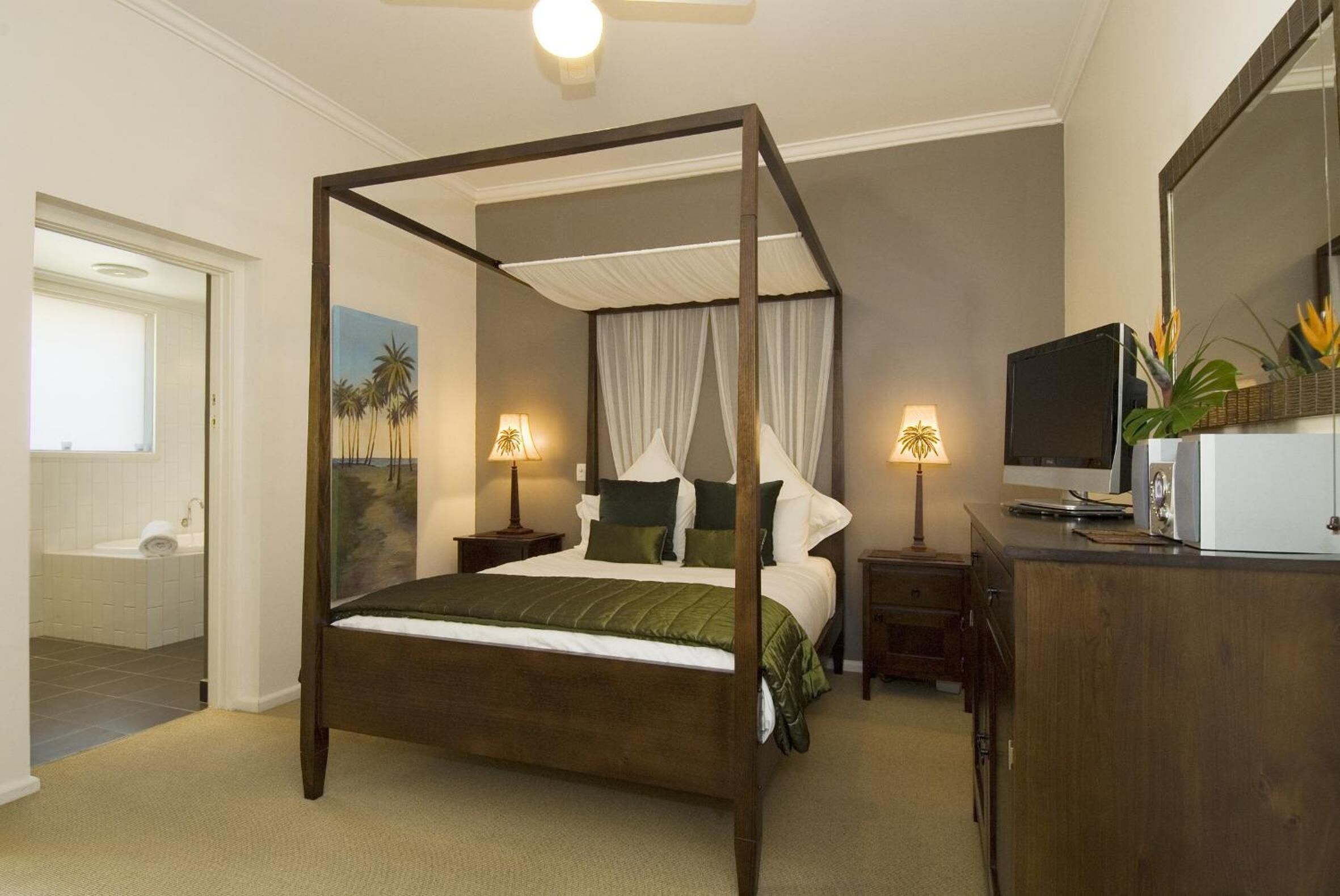 A four-poster bed with white linens, two bedside tables with lamps, a television on a wooden cabinet, and a bathroom visible through an open door.