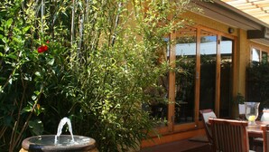 A wooden deck with a table and chairs, surrounded by lush greenery and a large pot with a water feature. - Plantation House (Rye)