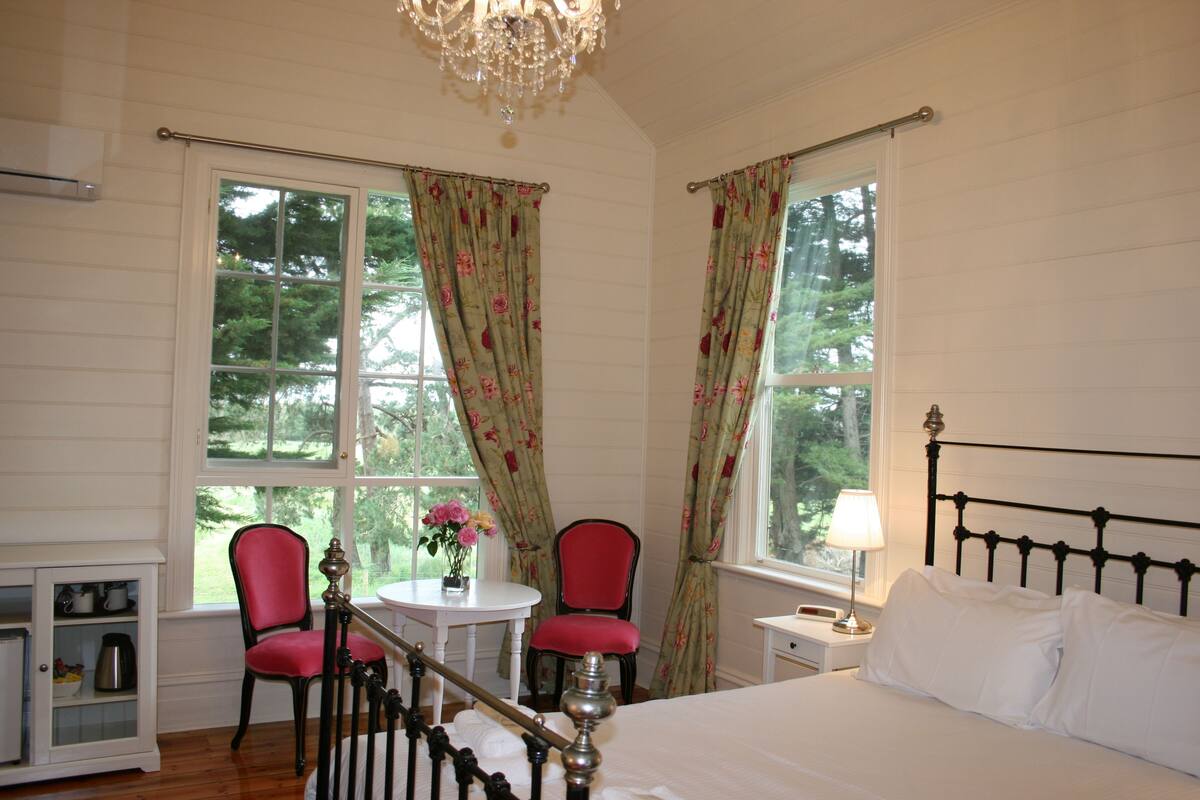 A bedroom with a bed, two chairs, a small table with flowers, a nightstand with a lamp, and a chandelier.