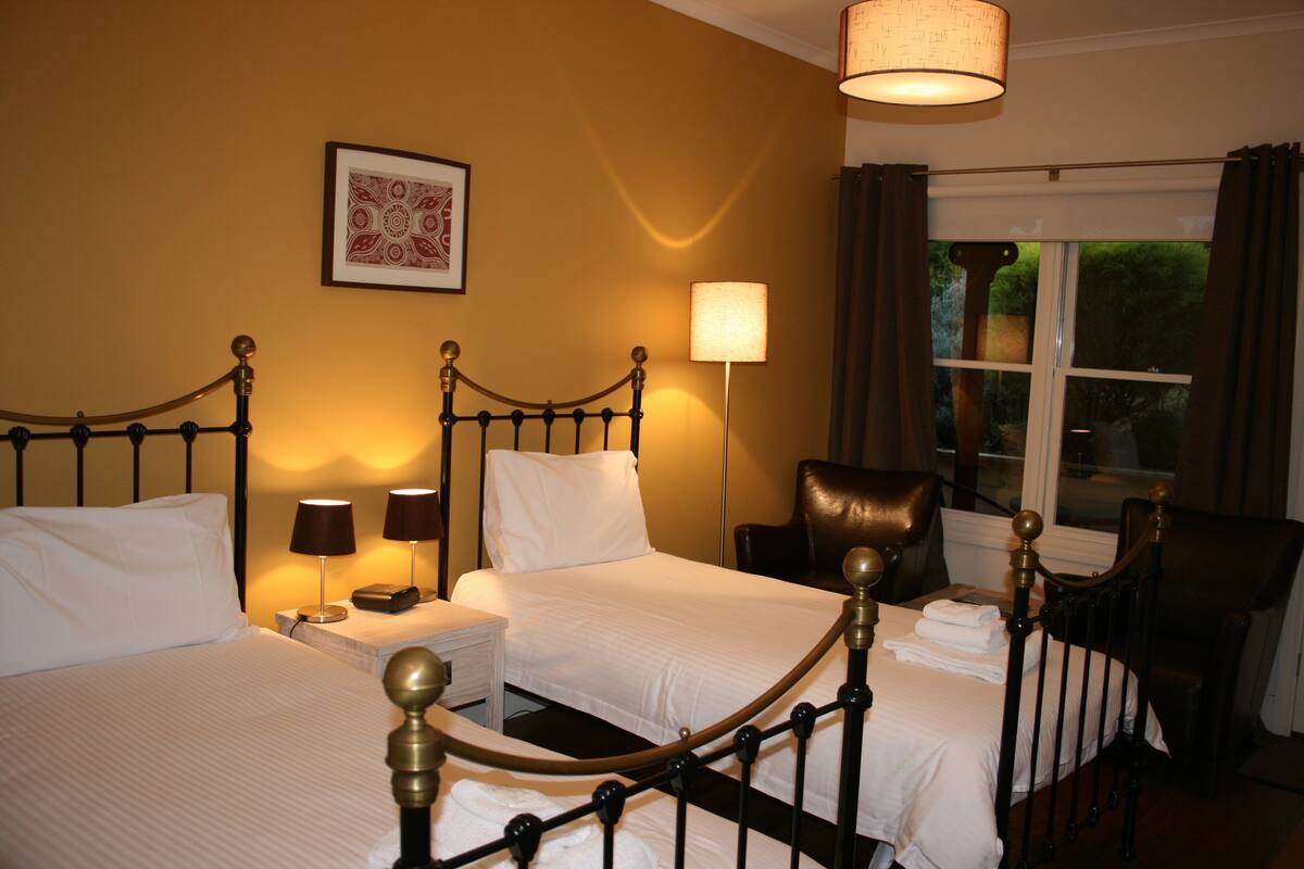 A hotel room with two beds, a chair, a lamp, a window with curtains, and a wall-mounted picture.