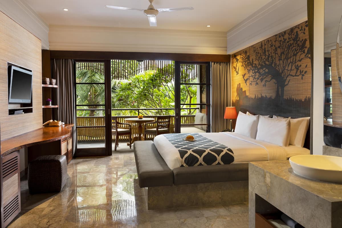 Habitación de hotel con cama, escritorio, televisor, balcón con vistas a zonas verdes y un cuadro en la pared.