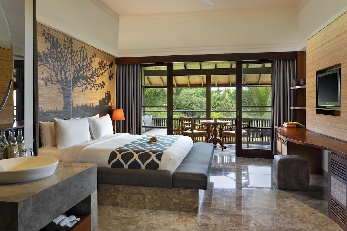 Habitación de hotel con cama, escritorio con televisor, baño con lavabo y ventana grande con vistas a zonas verdes.