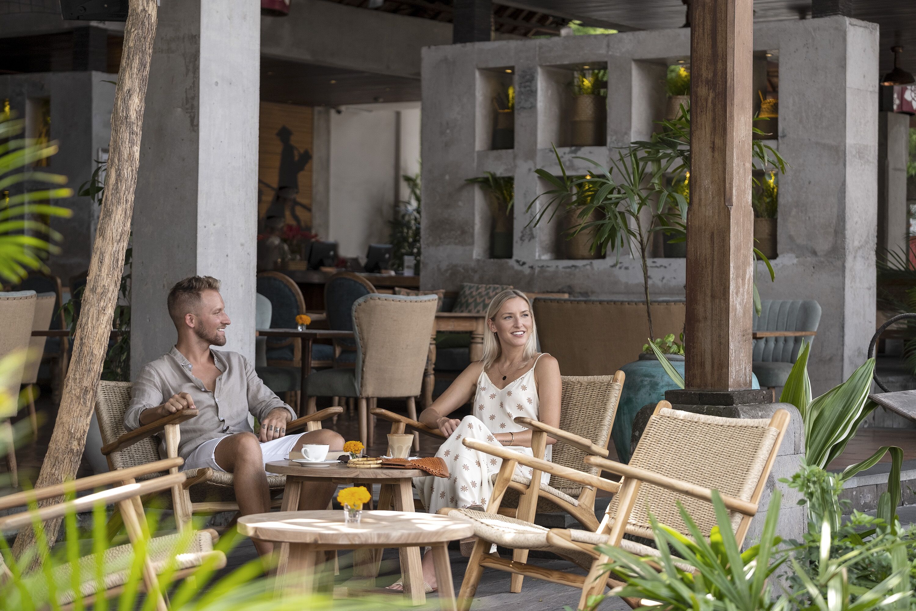 A couple seated at a round table in an outdoor cafe area with wicker chairs and potted plants.