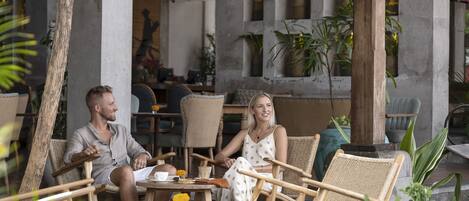A couple seated at a round table in an outdoor cafe area with wicker chairs and potted plants.