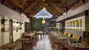 A restaurant with a high ceiling and natural light, featuring wooden tables and chairs, a sofa, and a view of greenery outside.