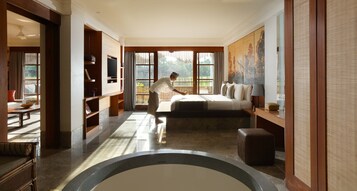 A spacious bedroom with a large bed, a sitting area with a sofa and a coffee table, a television, and a view of the outdoors through large windows.