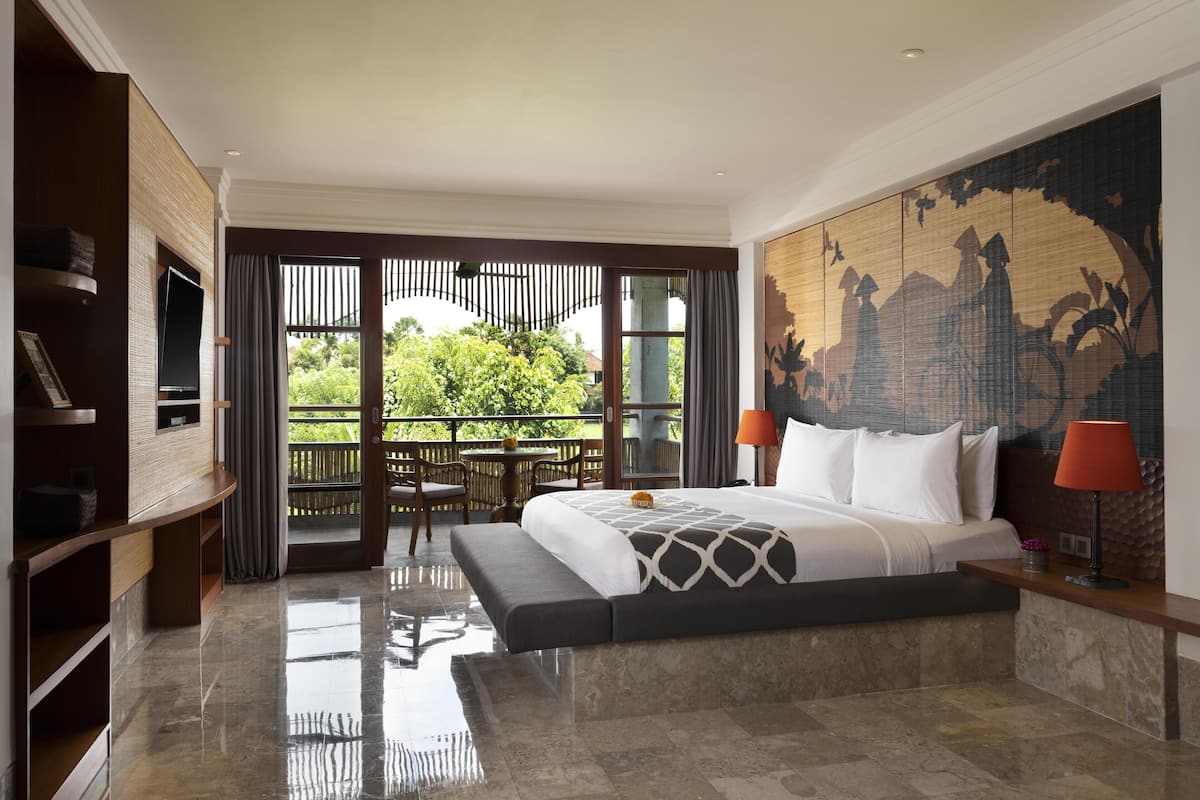 Un dormitorio amplio con una cama grande, un televisor montado en un panel de madera, un balcón con vistas a zonas verdes y un mural decorativo en la pared.