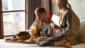 Una pareja sentada en una cama con artículos de desayuno, incluyendo una cesta de pan y un vaso de zumo de naranja.
