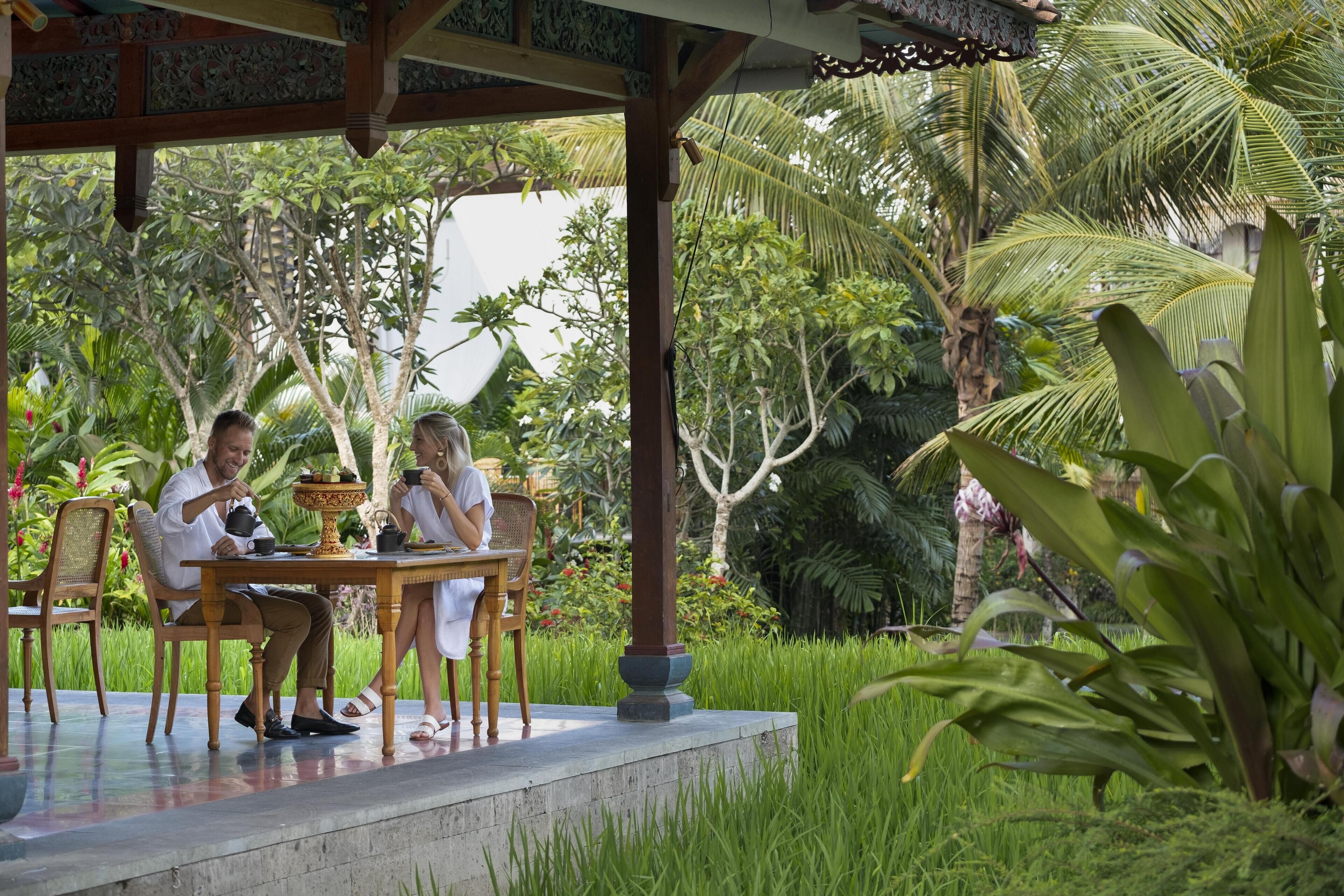 A covered outdoor seating area with a table and chairs, surrounded by lush greenery and palm trees.