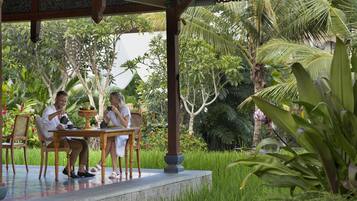 A covered outdoor seating area with a table and chairs, surrounded by lush greenery and palm trees.