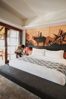 Habitación de hotel con una cama grande, cabecera de madera con un mural y una mesita de noche con lámpara, además de vistas al exterior a través de una puerta corredera.
