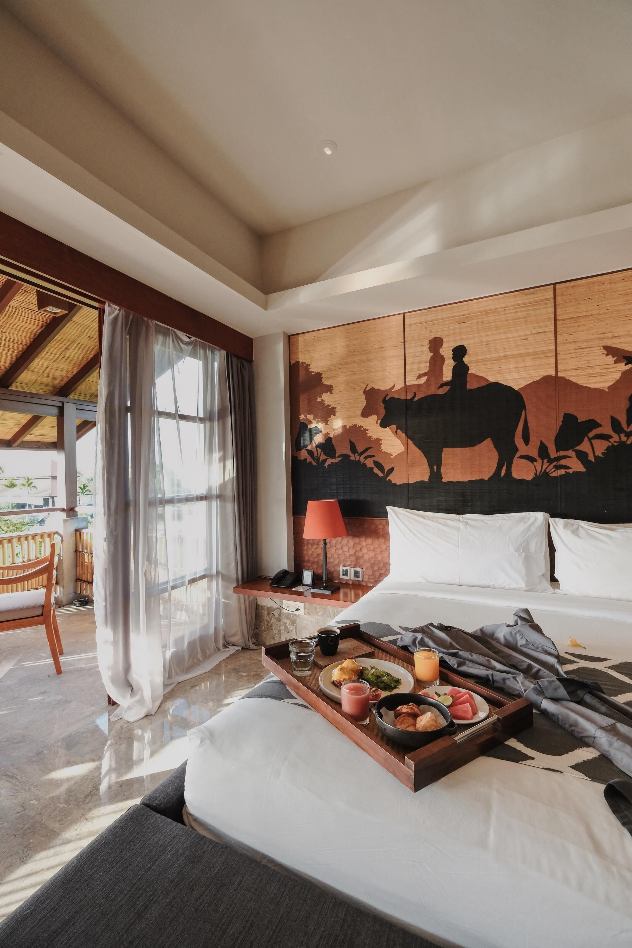 Een hotelkamer met een bed, een dienblad met ontbijt, een houten hoofdbord met een muurschildering, een lamp en uitzicht op het omliggende landschap.