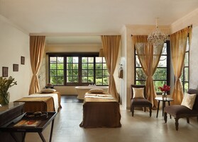 Quarto de hotel com duas camas, um espaço de estar com uma poltrona e uma mesa, uma banheira e janelas amplas com cortinas.