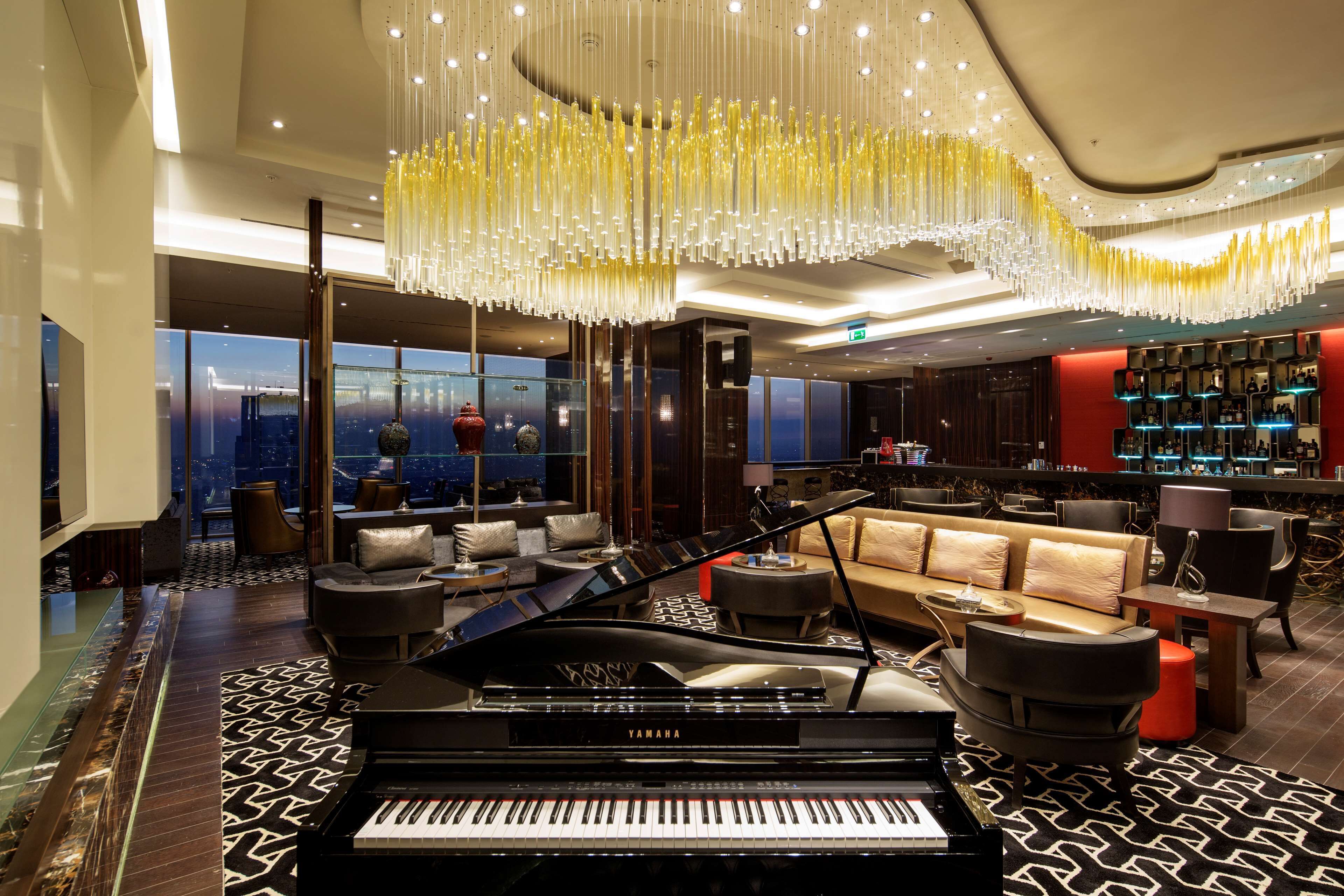 A grand piano in a spacious lobby with a modern chandelier, seating area, and a bar.