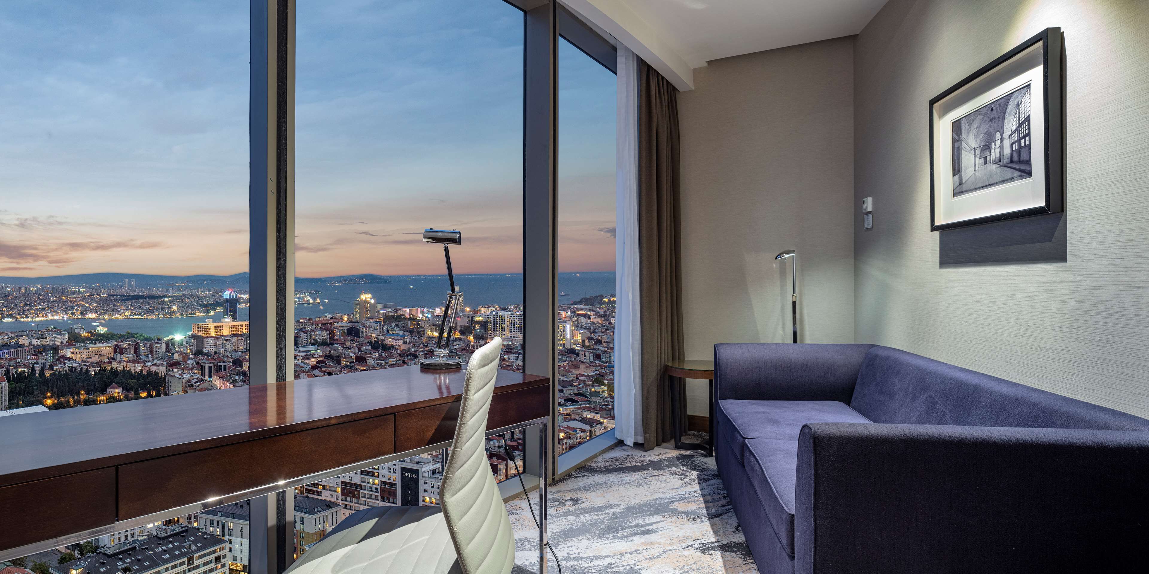 A modern hotel room with a large window offering a city view, a comfortable sofa, and a framed artwork on the wall.