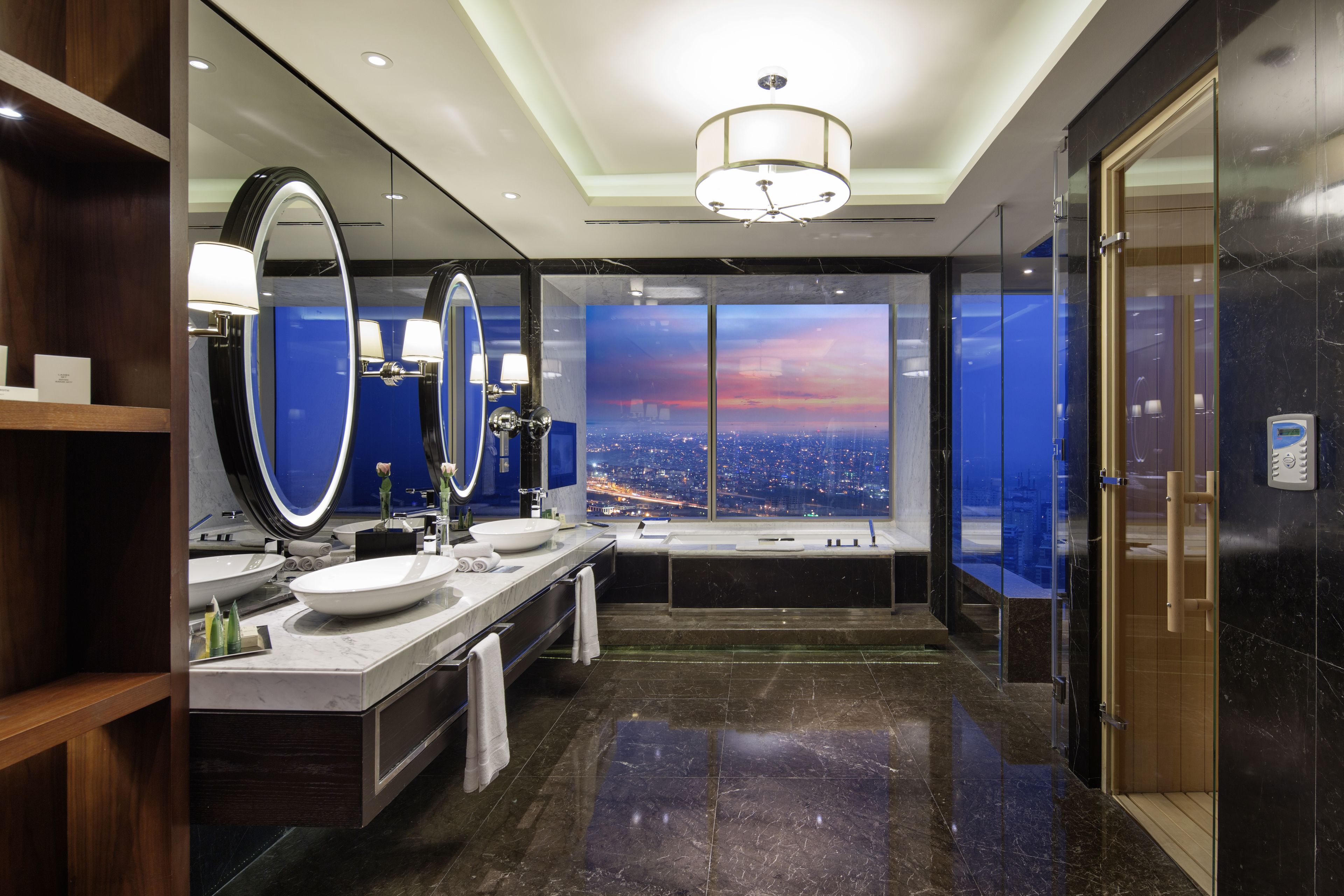 A modern bathroom with a large mirror, two sinks, a bathtub, and a view of a cityscape at sunset.