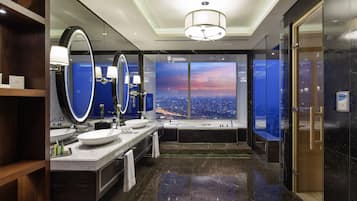 Une salle de bain moderne avec un grand miroir, deux vasques, une baignoire et une vue sur le paysage urbain au coucher du soleil.