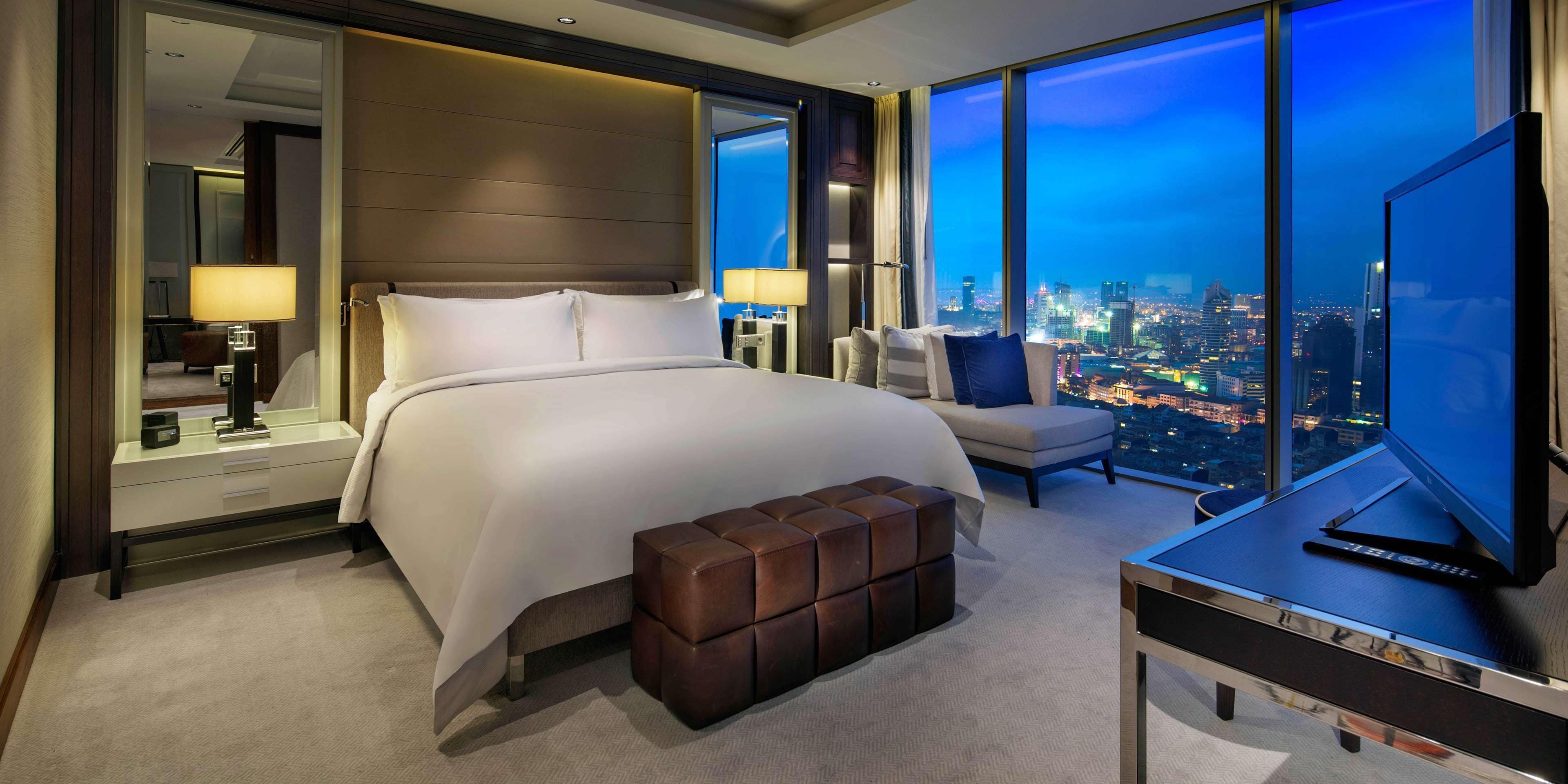 A modern hotel room with a large bed, a seating area, a desk with a TV, and a city view.