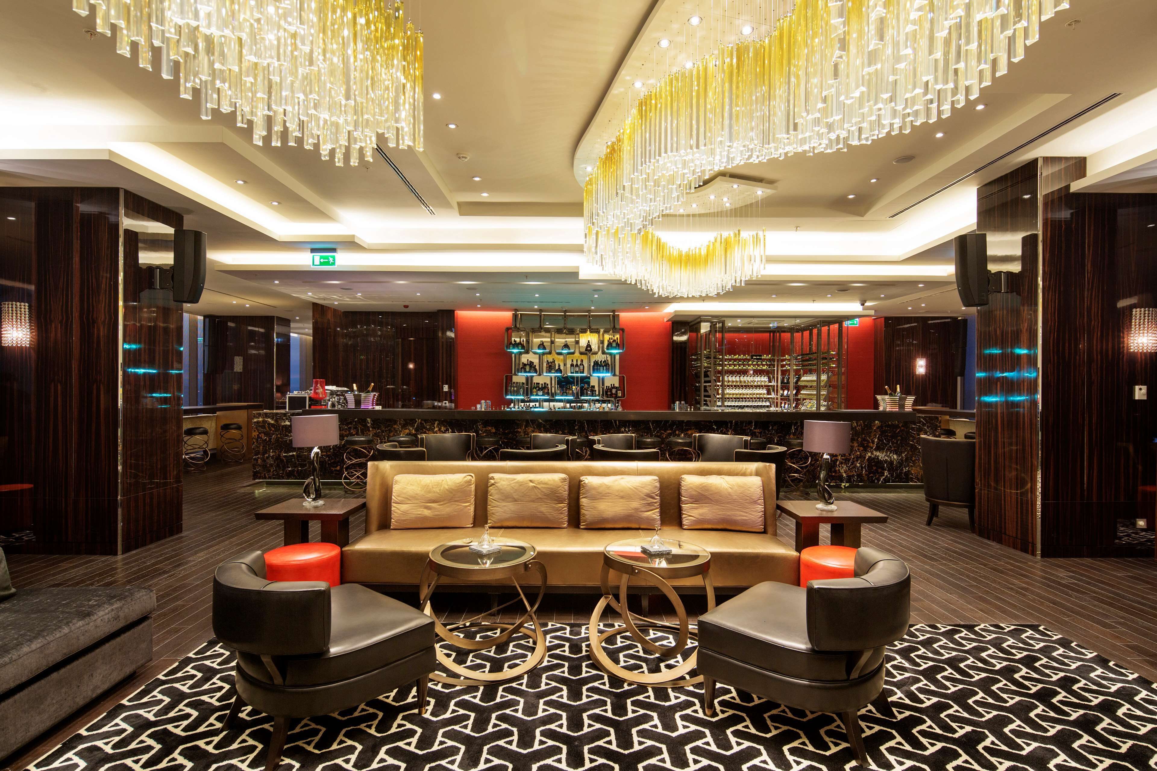 A modern lobby with a central seating area, a bar with a red accent wall, and a patterned carpet.
