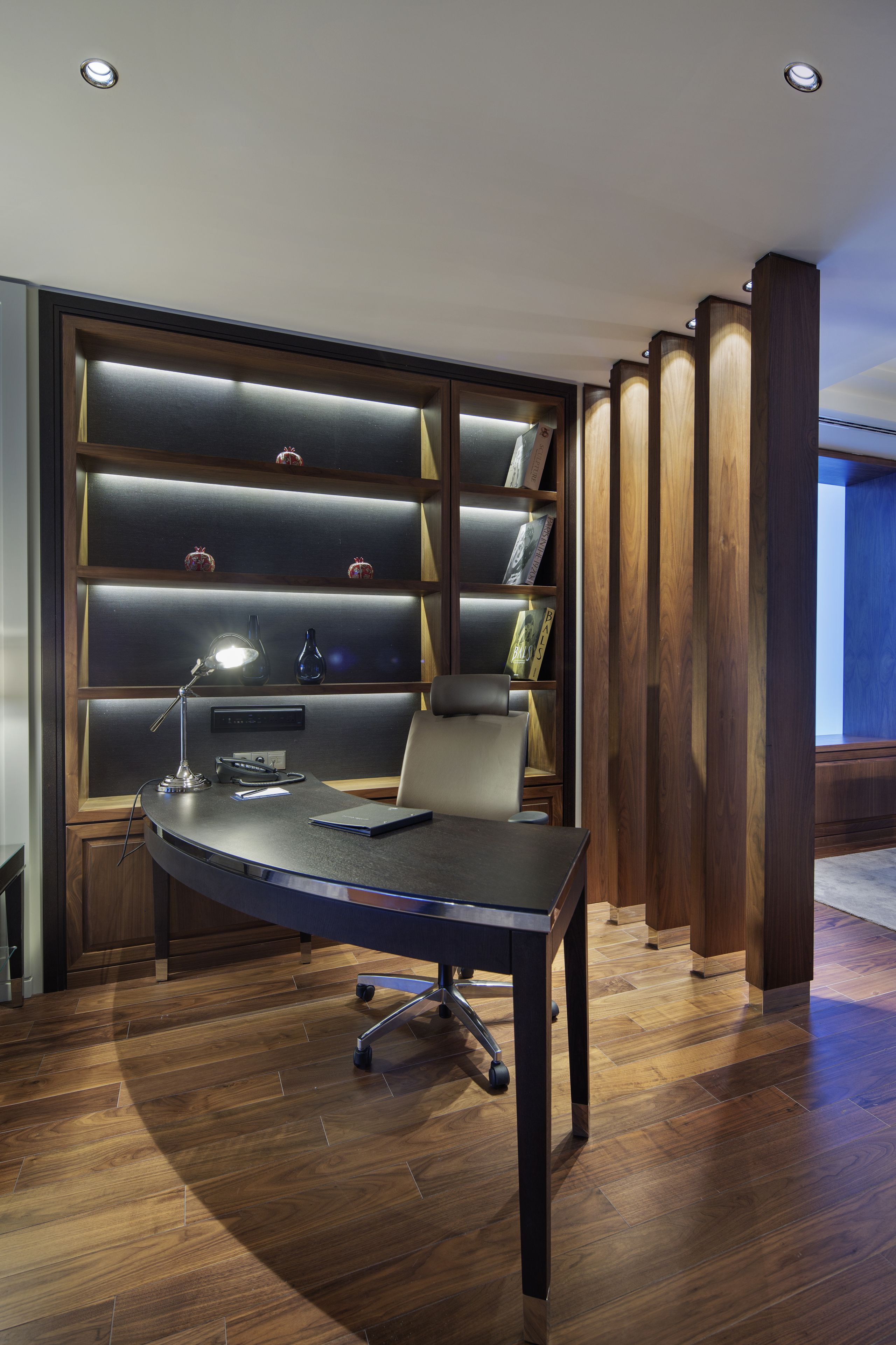 A modern office space with a curved desk, a chair, a desk lamp, and a bookshelf with illuminated shelves.