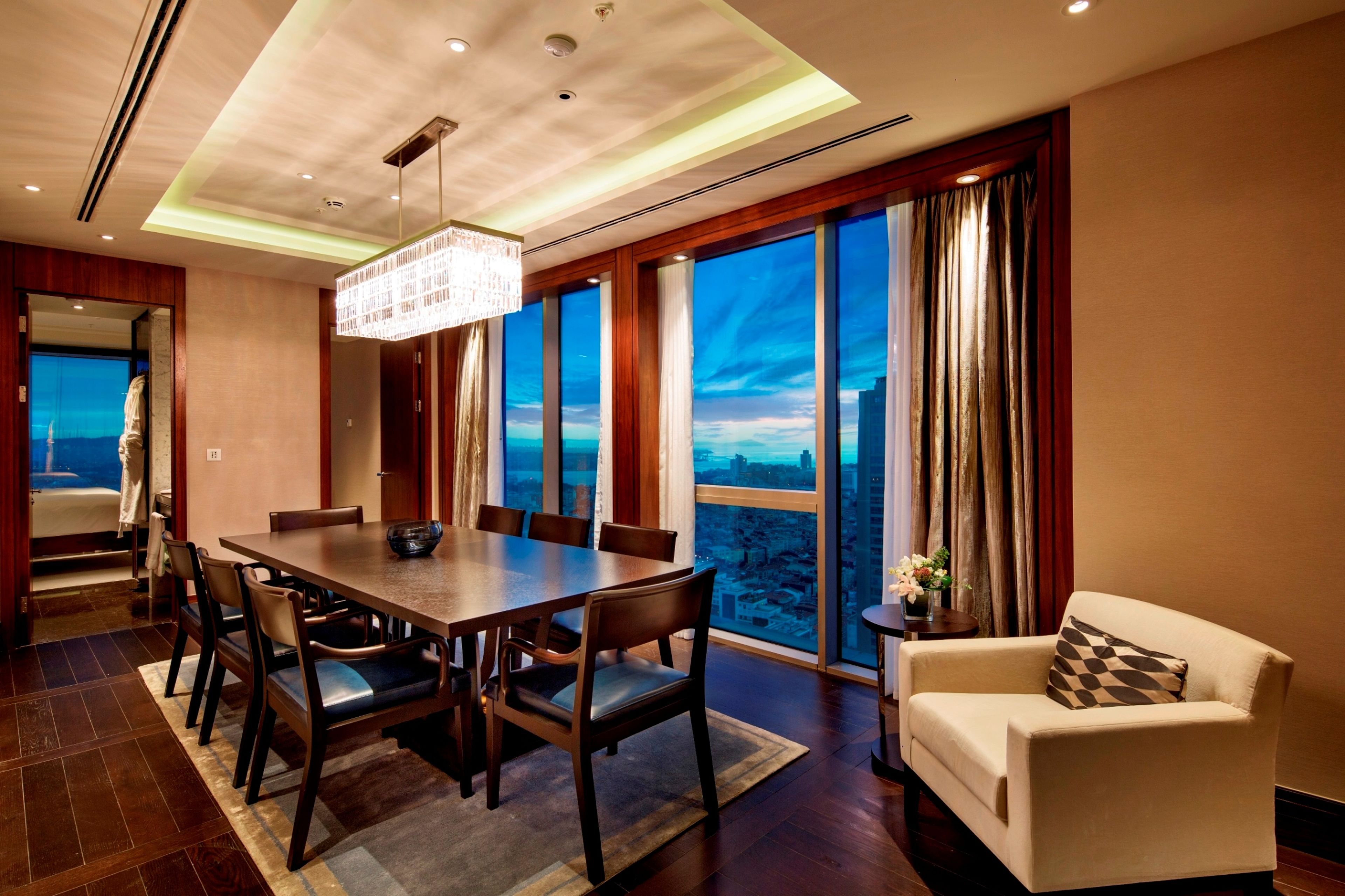 A modern dining area with a large table and chairs, a chandelier, and a view of the cityscape through large windows.