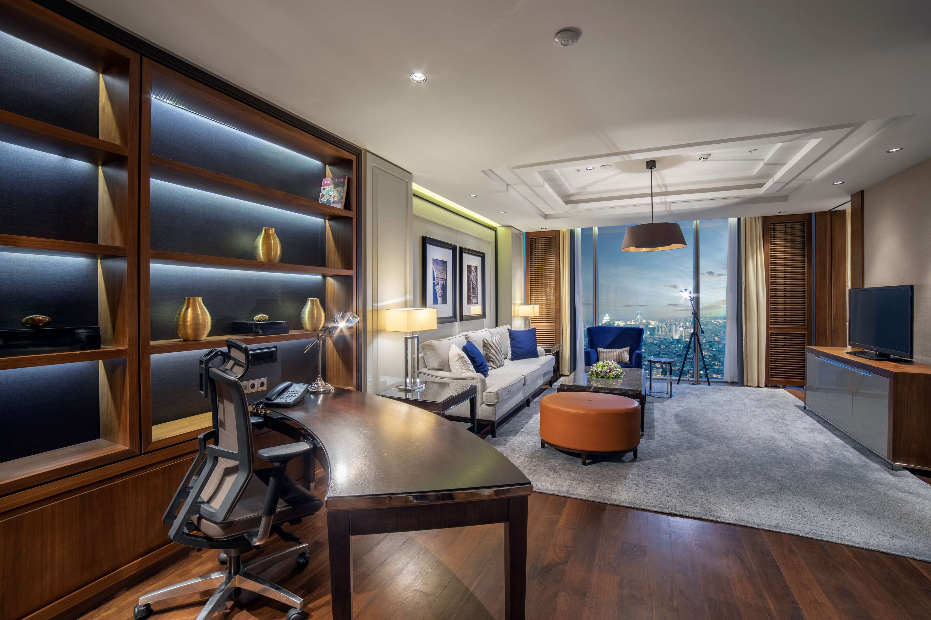 A modern hotel room with a wooden desk, a sofa, a TV, and a large window offering a city view.