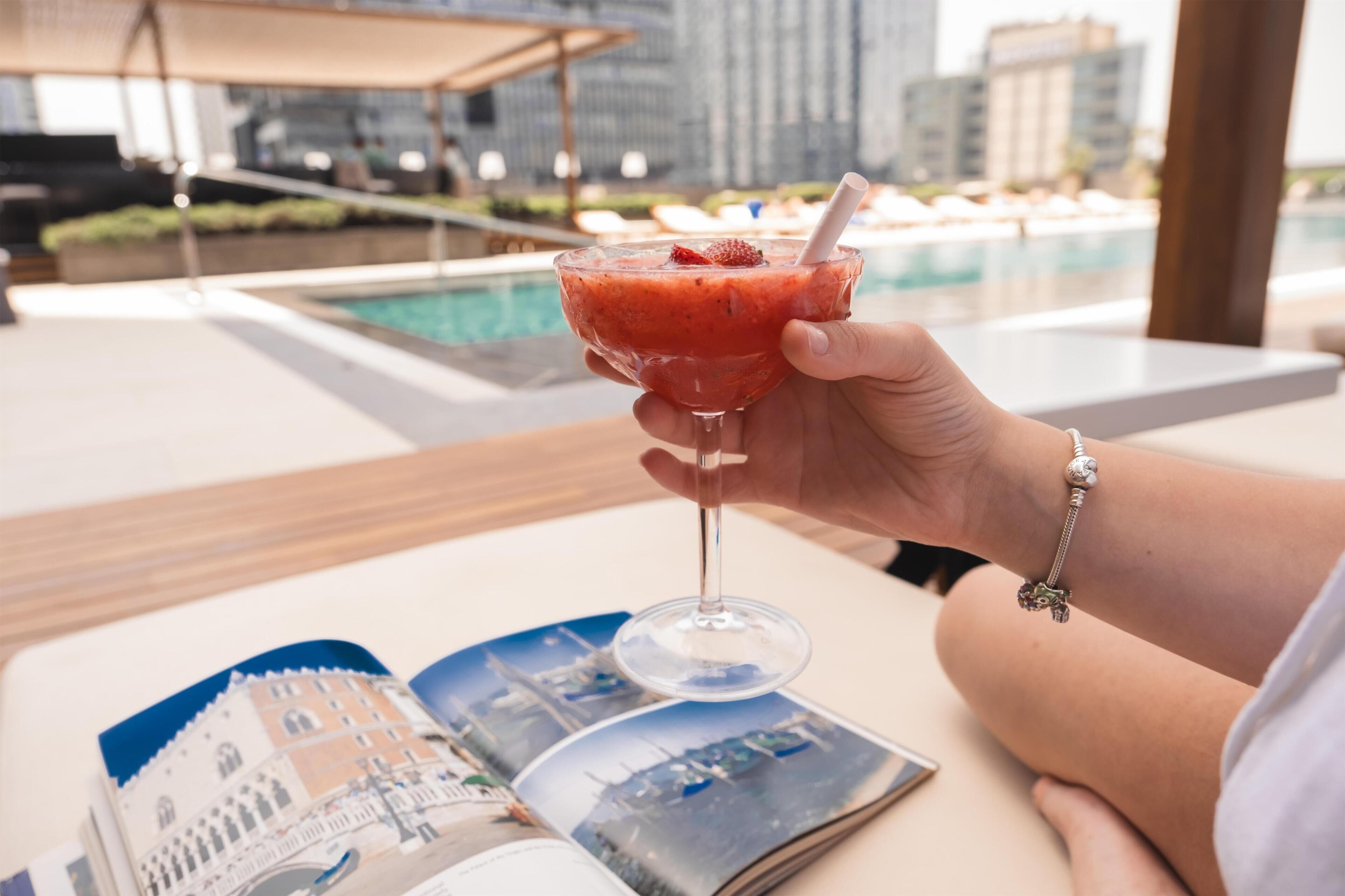 Una mano sosteniendo un vaso con una bebida roja, una piscina y una revista sobre una mesa.