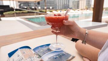 Una mano sosteniendo un vaso con una bebida roja, una piscina y una revista sobre una mesa.
