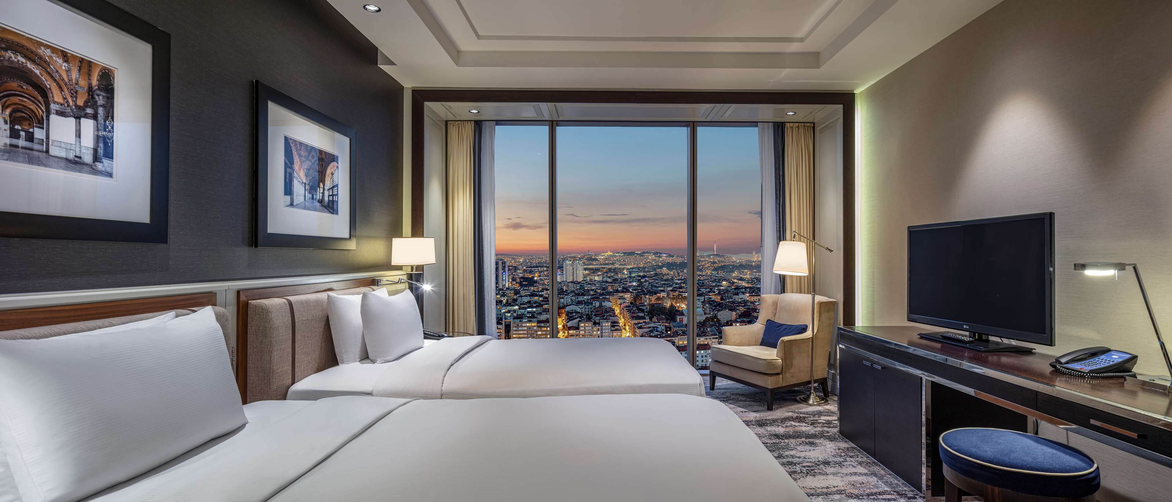 Habitación de hotel con dos camas, un escritorio, un televisor y un ventanal con vistas a la ciudad.