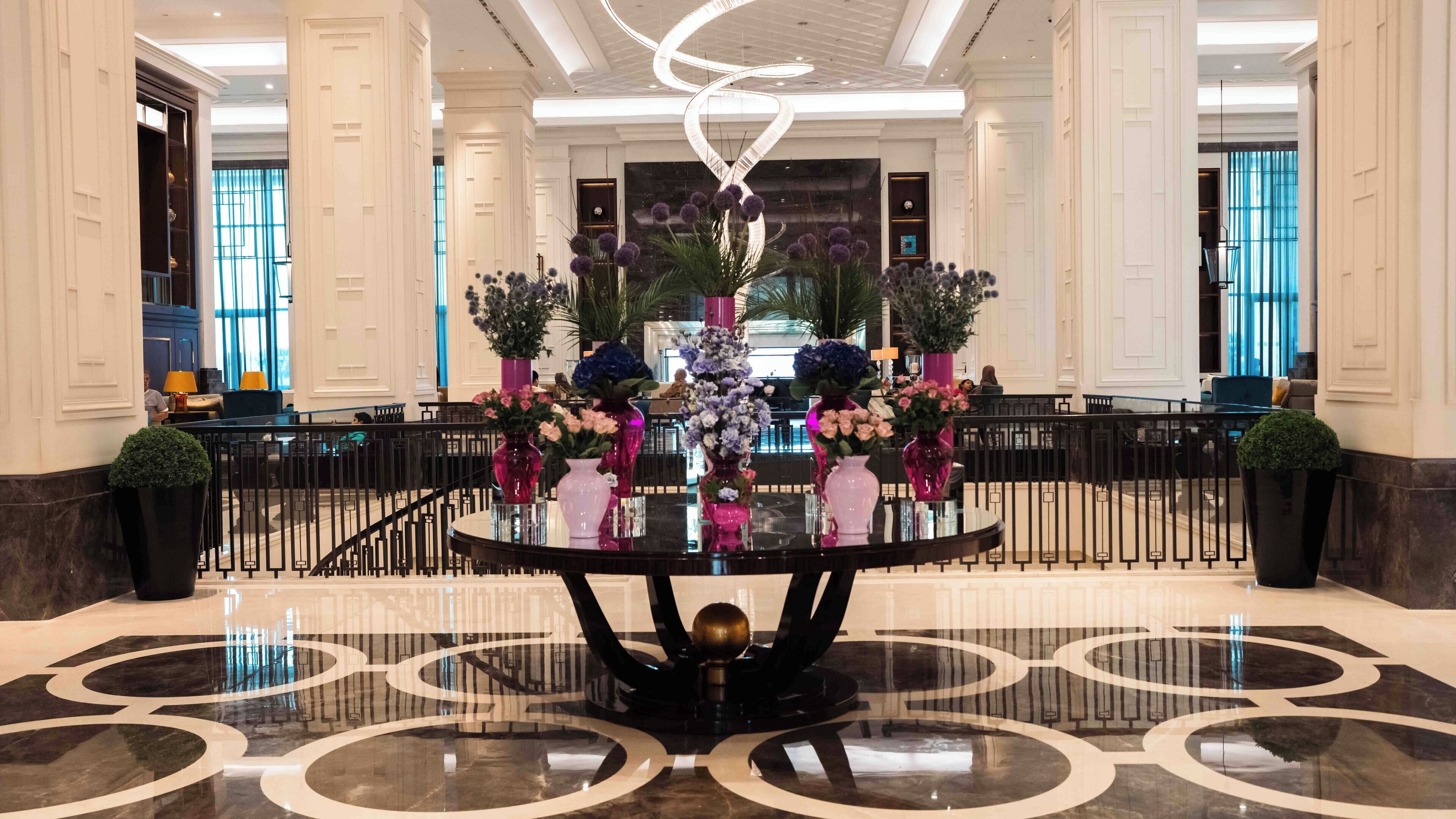 A lobby with a round table surrounded by vases of flowers, a spiral light fixture, and a marble floor with circular patterns.