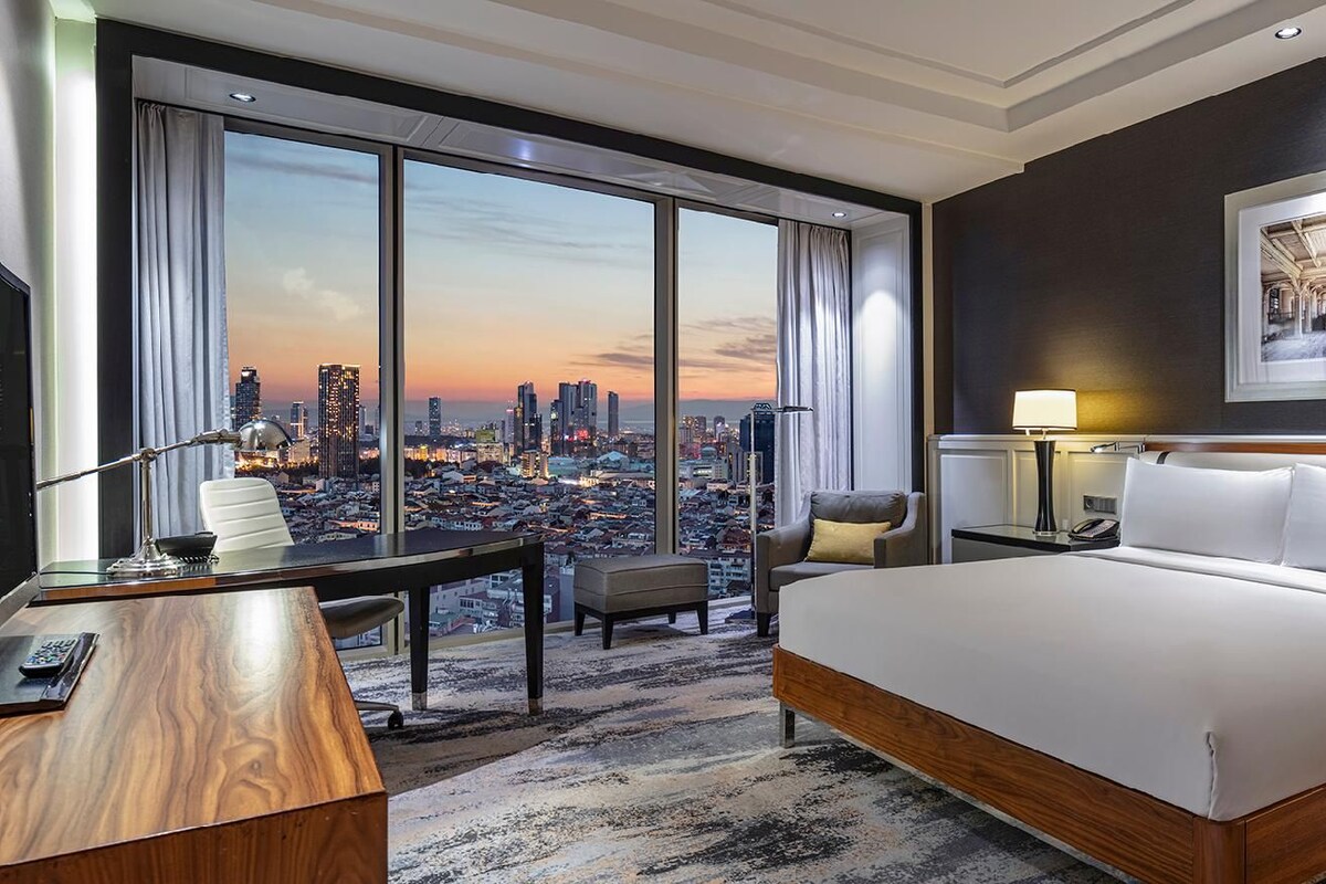 A hotel room with a large bed, a desk, a chair, a TV, and a city view.