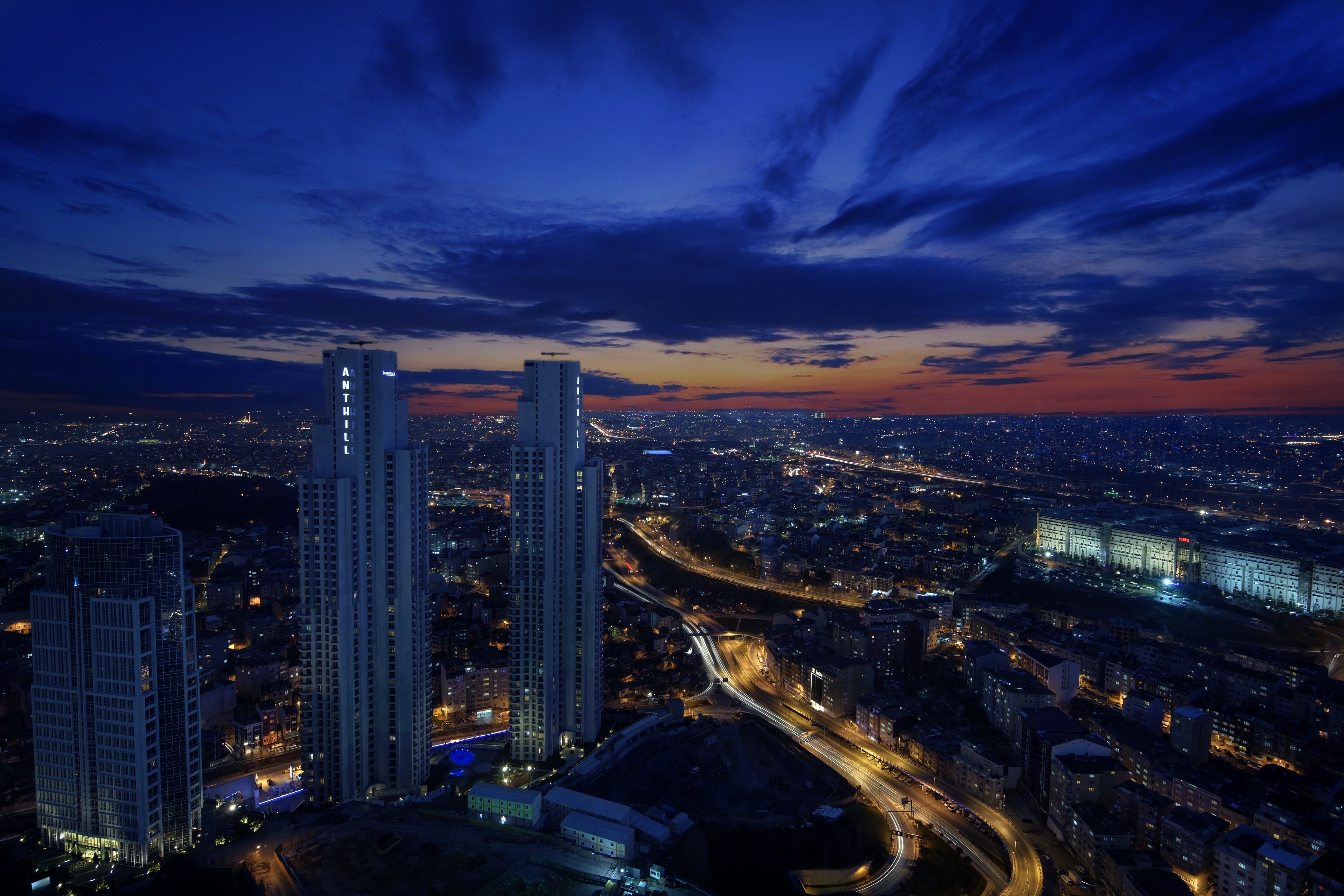 Une vue nocturne d’une ville, avec des gratte-ciel, une autoroute et un grand bâtiment dont les fenêtres sont illuminées.