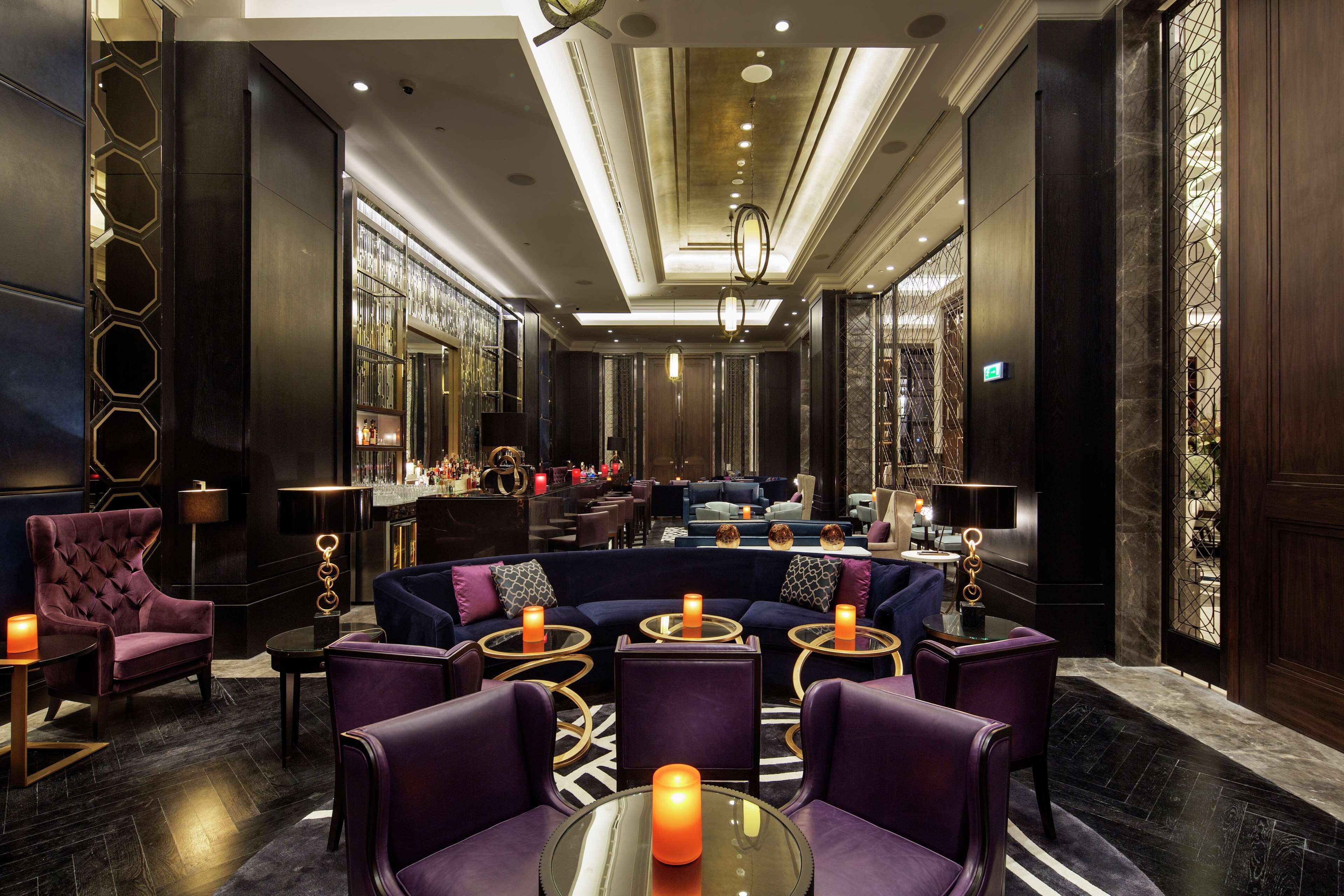 A luxurious lobby with a dark color scheme, featuring a seating area with purple upholstered chairs and tables, a bar with a glass railing, and a chandelier.