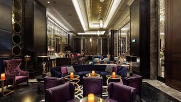 A luxurious lobby with a dark color scheme, featuring a seating area with purple upholstered chairs and tables, a bar with a glass railing, and a chandelier.