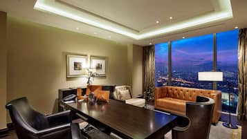 A modern hotel room with a large desk, leather chairs, a sofa, and a large window offering a city view.