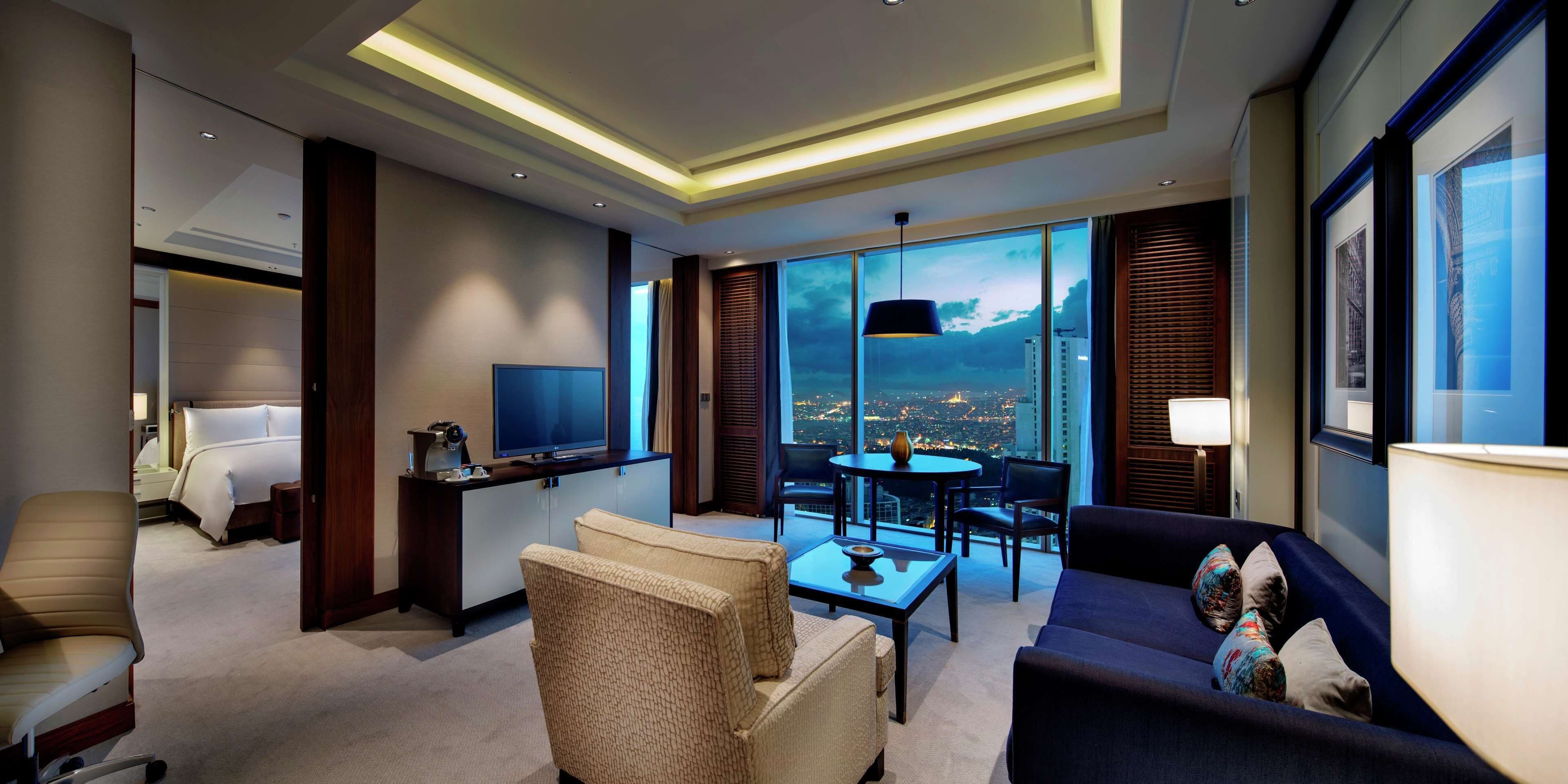 A modern hotel room with a large bed, a TV, a sofa, a coffee table, and a balcony with a city view.