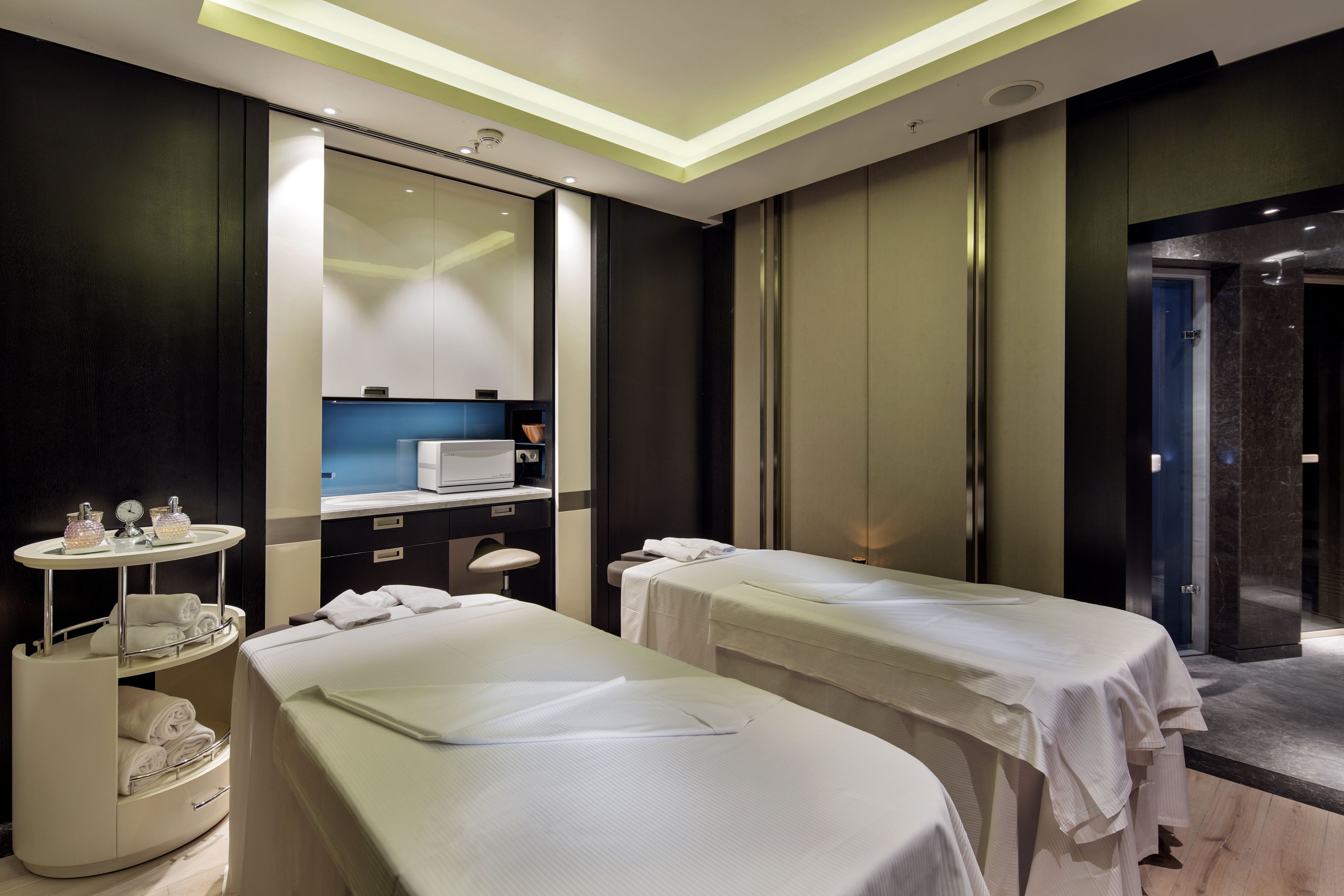 A modern spa room with two massage tables, a small vanity with a mirror, and a sink.