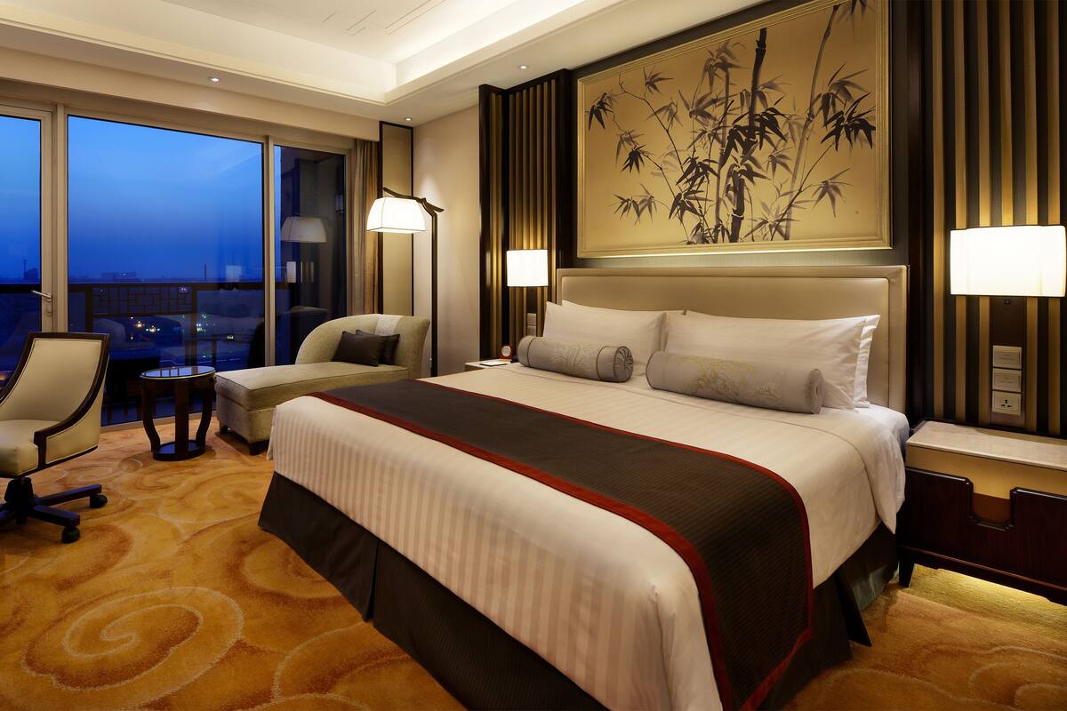 Une chambre d’hôtel avec un grand lit, un bureau, une chaise et une vue sur la ville au crépuscule.