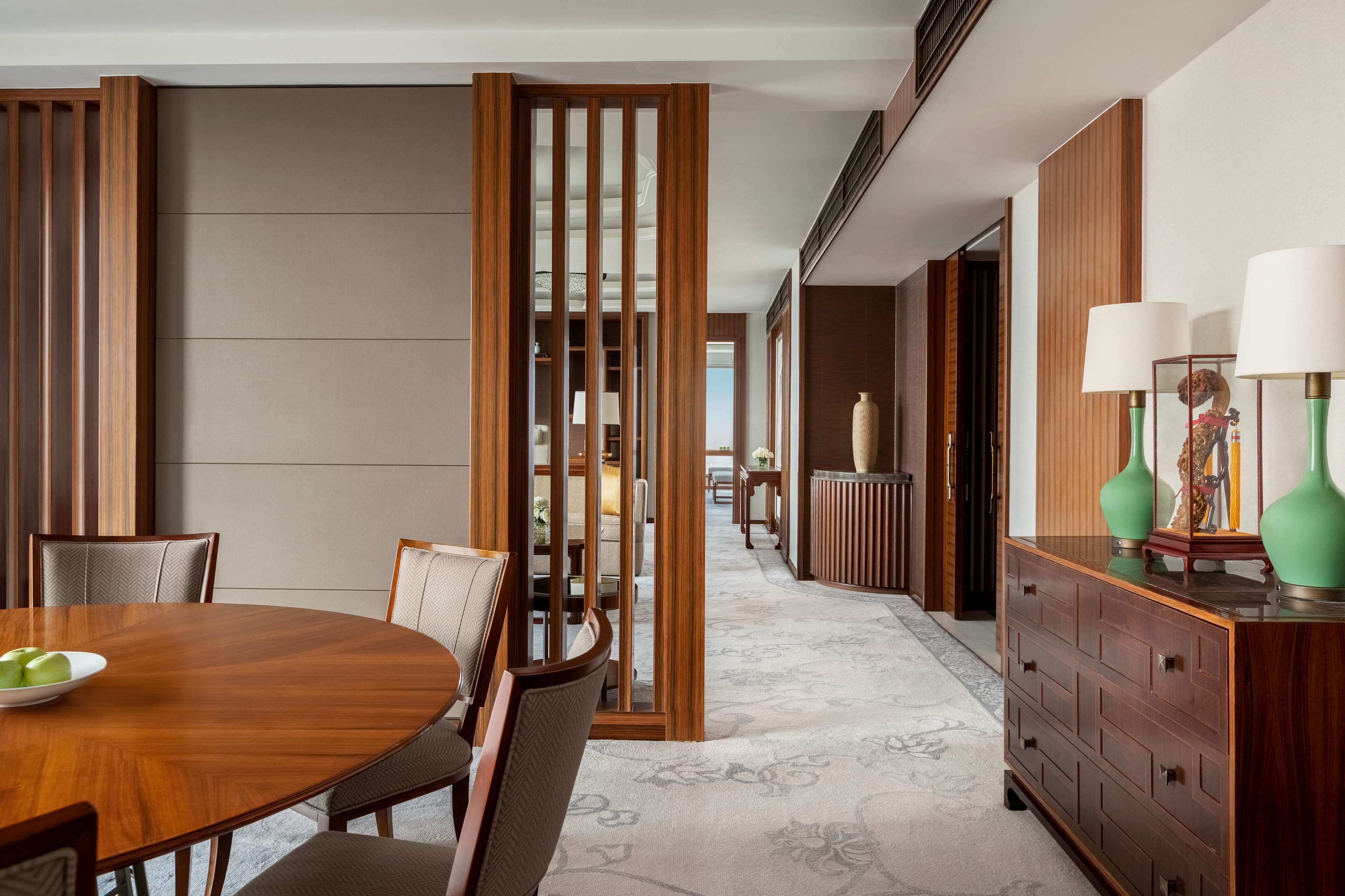 A modern hotel room with a wooden dining table, chairs, a large mirror, and a dresser with lamps.
