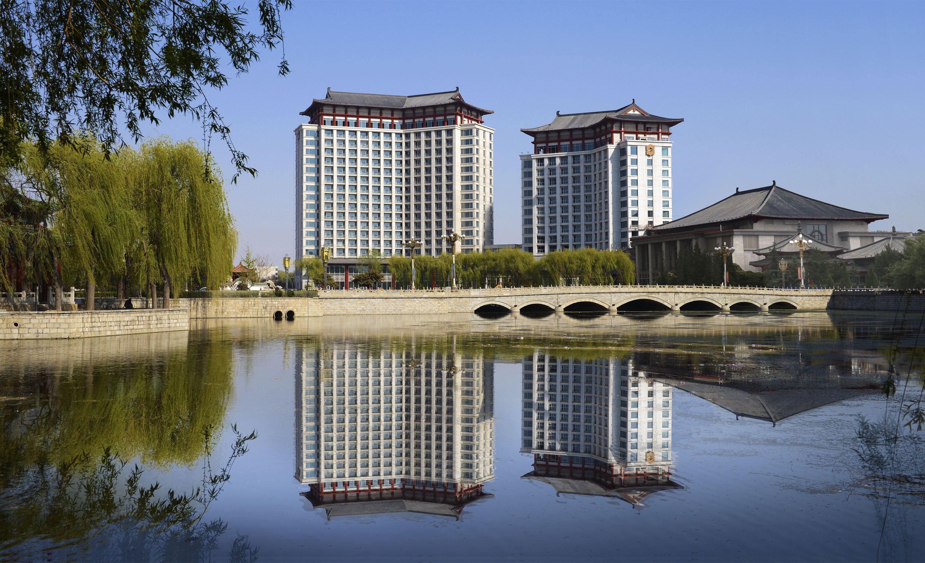 A traditional building with a modern high-rise in the background, a bridge over a calm water body, and trees on the left.