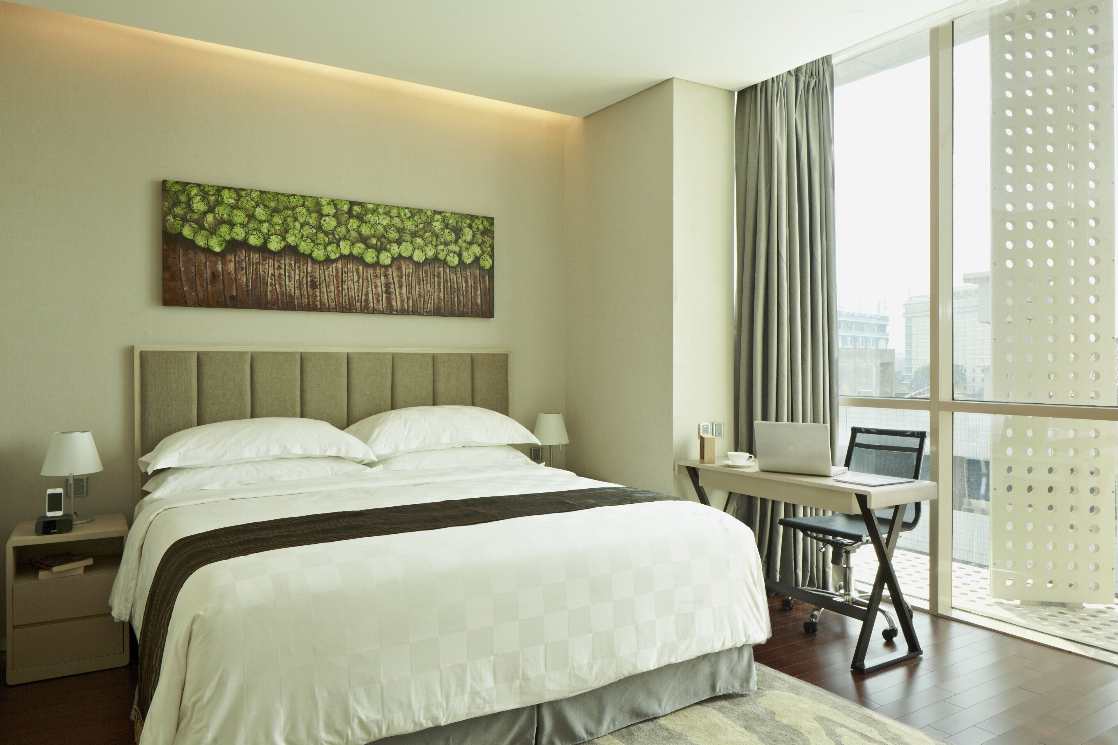 A hotel room with a bed, a desk with a laptop, a nightstand with a lamp, and a large window with a city view.