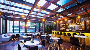 An indoor bar area with wooden flooring, round tables, black chairs, a bar counter, and large windows offering views of the outside. - Fraser Residence Menteng Jakarta (Jakarta)