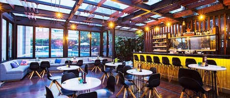 An indoor bar area with wooden flooring, round tables, black chairs, a bar counter, and large windows offering views of the outside.