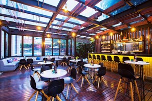 An indoor bar area with wooden flooring, round tables, black chairs, a bar counter, and large windows offering views of the outside. - Fraser Residence Menteng Jakarta (Jakarta)