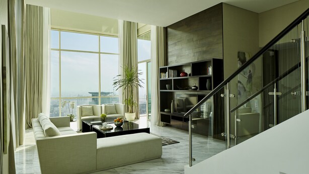 A modern living room with a sofa, coffee table, and a bookshelf. There is a staircase with glass railings leading to an upper floor. - Fraser Residence Menteng Jakarta (Jakarta)