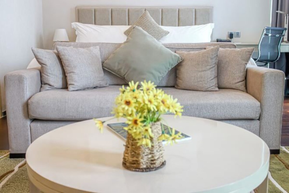 A neutral-toned living area with a sofa, a round coffee table with a woven basket and flowers, and a bed with a headboard in the background.