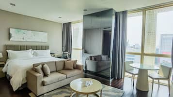 Quarto de hotel com cama, sofá, mesa redonda e amplas janelas com vista para a cidade.